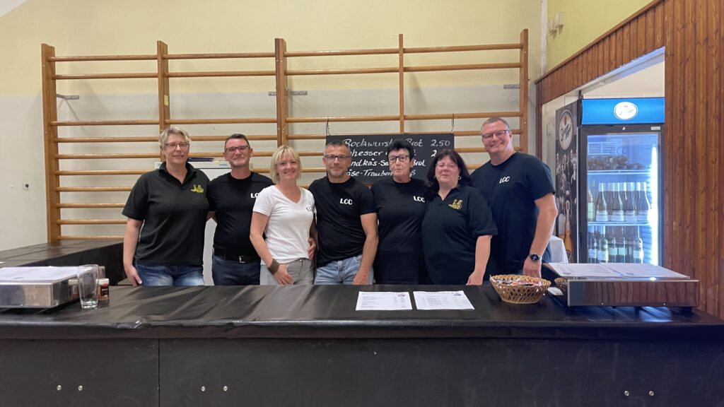 Die Crew des LCC vor ihrer Bar in der Turnhalle, 4 Frauen und drei Männer in Shirts des LCC sind zu sehen, dahinter ein Preisschild und ein Kühlschrank